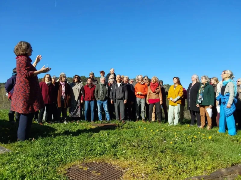 Le chœur chante en plein air sur l'herbe, la cheffe de chœur dirige à gauche, sous un grand ciel bleu